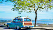 Los Angeles, California /USA - 06 30 2020: Old classic retro car with surfboards and beach tool on the roof of the car, at shade underneath a beautiful green-leafed tree, some place along the coast.
