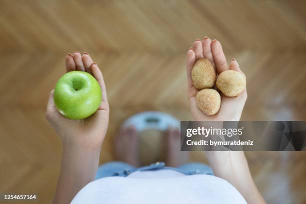 young woman holding green apple and sweet on a weigh scale at home - fat loss stock pictures, royalty-free photos & images