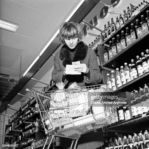 In der Spirituosenabteilung in einem Supermarkt in Hamburg prüft eine Kundin ihren Einkaufszettel, Deutschland Anfang 1960er Jahre.