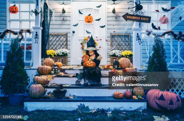 kleines mädchen in hexenkostüm sitzt auf der treppe vor dem haus und hält jack-o-lantern kürbisse auf halloween-trick oder behandeln - halloween candy stock-fotos und bilder