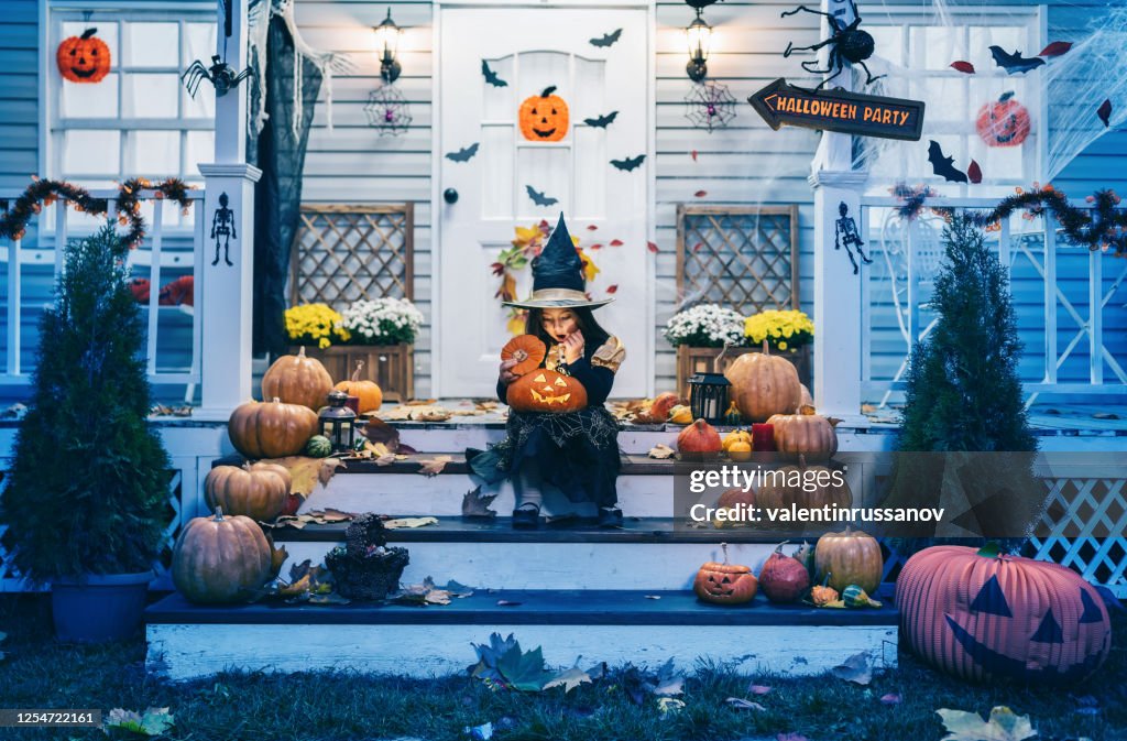 Little girl in witch costume sitting on the stairs in front of the house and holding Jack-o-Lantern Pumpkins on Halloween trick or treat