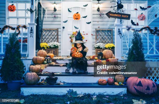 little girl in witch costume sitting on the stairs in front of the house and holding jack-o-lantern pumpkins on halloween trick or treat - jack o lantern imagens e fotografias de stock