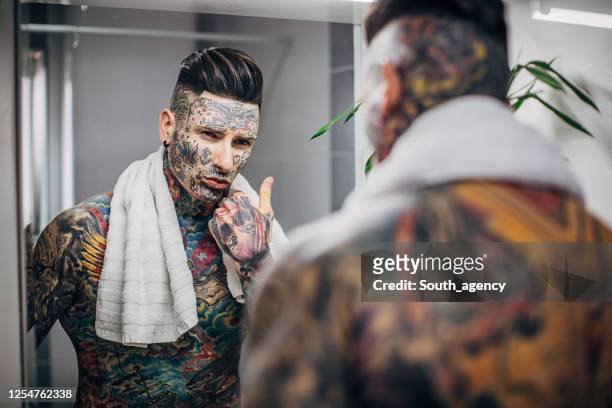 one man with whole body covered in tattoos looking at his face in the bathroom mirror - handsome tattoo stock pictures, royalty-free photos & images