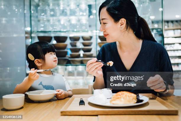 young asian mother and cute little daughter enjoying dessert in a cafe. they are looking at each other and smiling happily - asian family cafe stockfoto's en -beelden