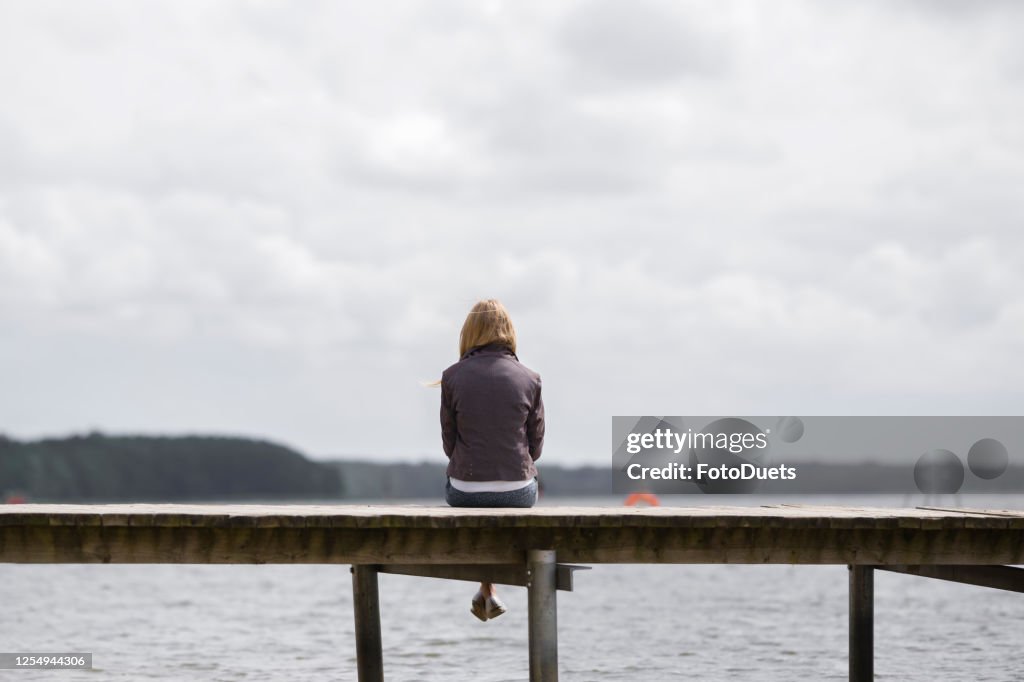 One young woman sitting alone on edge of footbridge and staring at lake and cloudy sky in summer day. Thinking about life. Spending time alone in nature. Peaceful atmosphere. Back view.
