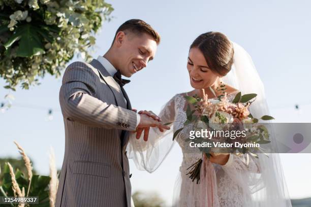 beautiful bride puts on a ring on her grooms finger - outdoor wedding ceremony vows stock pictures, royalty-free photos & images