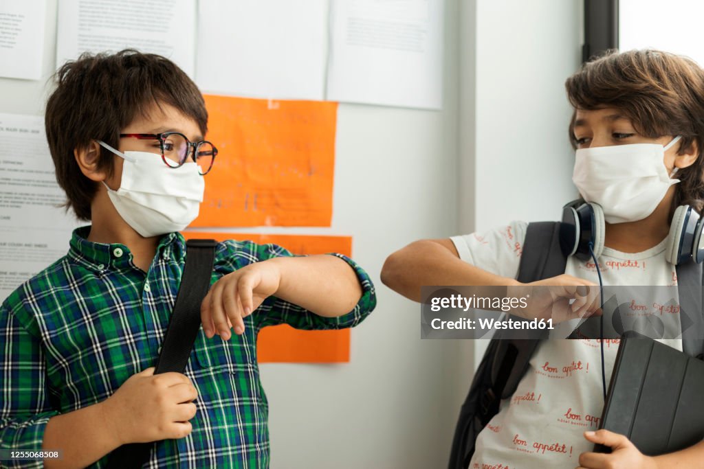 Boys wearing masks giving elbow bump while standing against wall in school
