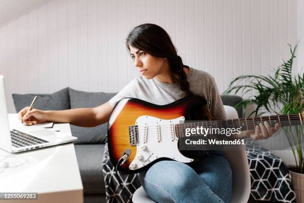 young woman writing while using laptop and electric guitar at home - writing instrument stock-fotos und bilder