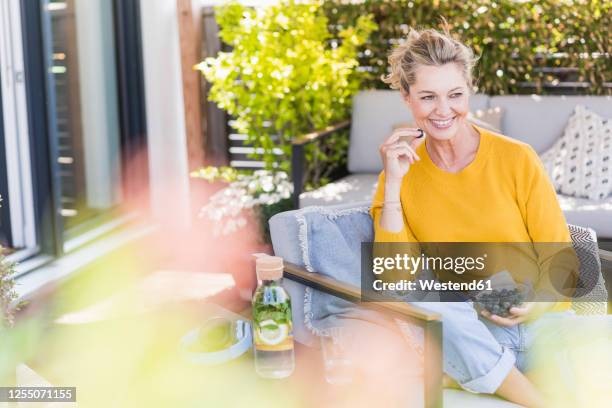portrait of happy mature woman sitting on terrace eating blueberries - blond women happy eating stockfoto's en -beelden