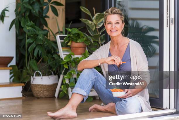 portrait of smiling mature woman sitting on the floor at open terrace door eating finger food - blond women happy eating stock pictures, royalty-free photos & images