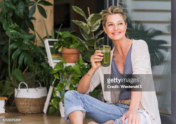 portrait of relaxed mature woman sitting at open terrace door with glass of green smoothie - open workouts foto e immagini stock