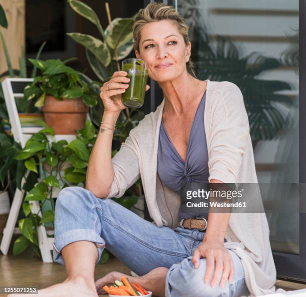 portrait of smiling mature woman sitting at open terrace door drinking green smoothie - mature woman smoothie stock-fotos und bilder