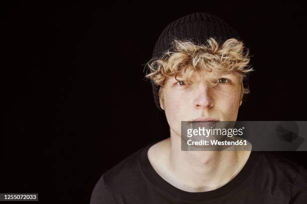 portrait of young man with curly blond hair wearing black cap against black background - kontrastreich stock-fotos und bilder
