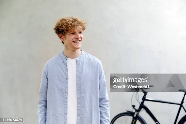 portrait of smiling young man with curly blond hair standing in front of grey wall - ventenne foto e immagini stock