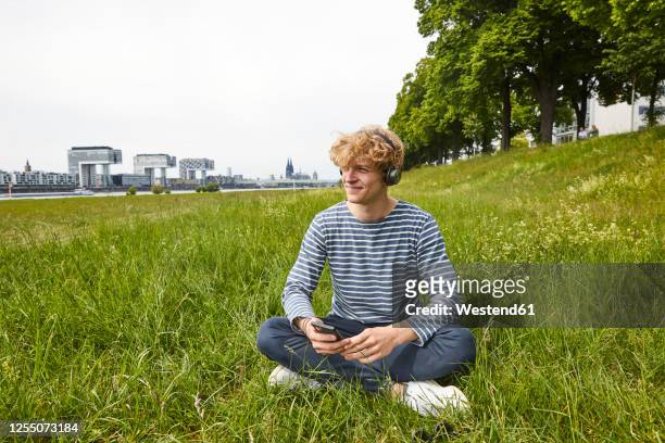 smiling young man sitting on a meadow listening music with headphones - mann lässig gras sitzen stock-fotos und bilder