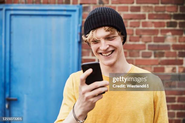 portrait of happy young man wearing cap looking at smartphone in front of brick wall - brick phone stock pictures, royalty-free photos & images