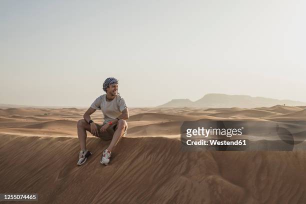 male tourist looking away while sitting on sand dunes in desert at dubai, united arab emirates - dubai tourist stock pictures, royalty-free photos & images