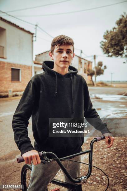 confident teenage boy standing with bicycle on street during rainy season - wet sweatshirt foto e immagini stock