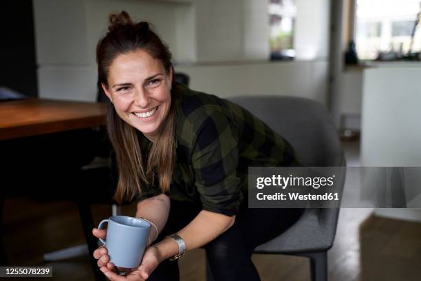 smiling woman holding coffee mug looking away while sitting on chair at home - one woman only 35-40 stock pictures, royalty-free photos & images