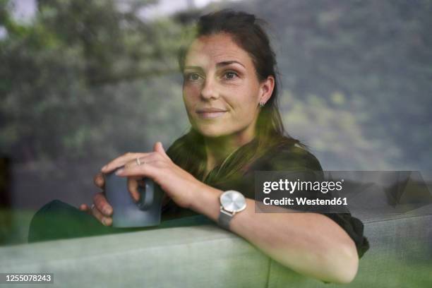 thoughtful woman holding coffee mug while relaxing on couch at home seen through window - woman window home stock pictures, royalty-free photos & images