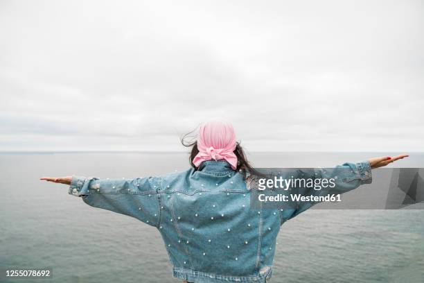 young female cancer survivor with arms outstretched looking at sea against cloudy sky - cancer survivor stockfoto's en -beelden