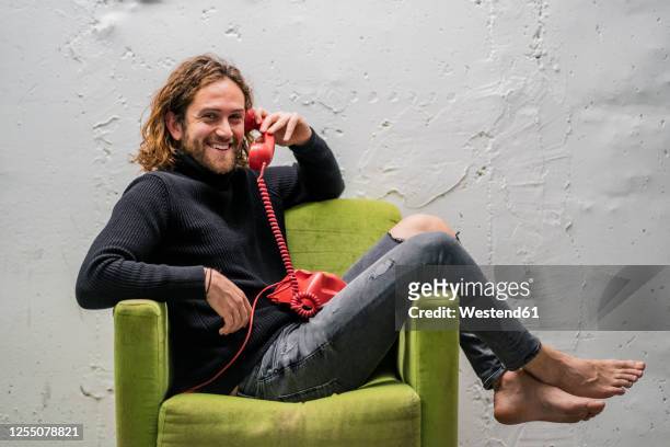 smiling man talking over telephone while relaxing on armchair against wall - telephone receiver fotografías e imágenes de stock