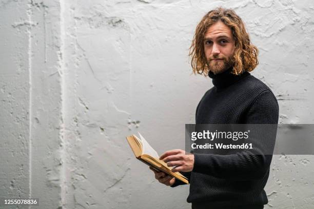 bearded young man holding book while standing against old wall at home - langes haar stock-fotos und bilder
