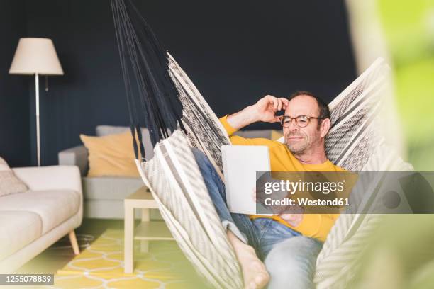 relaxed man holding digital tablet while sitting on hammock at home - hängematte stock-fotos und bilder