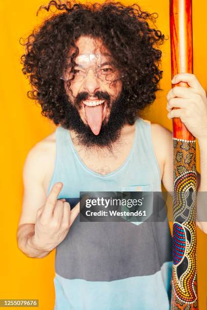 crazy man with curly hair holding didgeridoo while sticking out tongue against yellow background - didgeridoo stock pictures, royalty-free photos & images