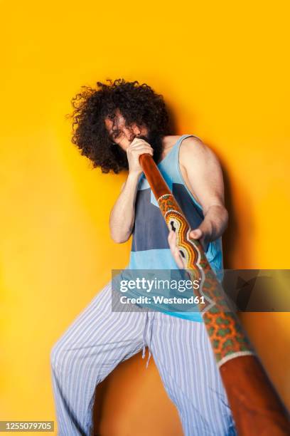 crazy mature man with curly hair playing didgeridoo while standing against yellow background - didgeridoo stock pictures, royalty-free photos & images