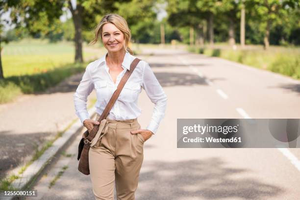 confident mature businesswoman carrying shoulder bag while walking on road - borsa monospalla foto e immagini stock