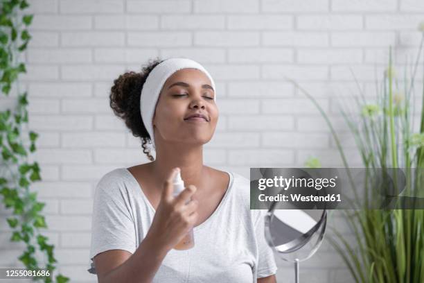 young woman with eyes closed spraying facial mist at home - botella para rociar fotografías e imágenes de stock