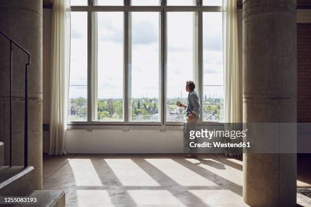 senior man looking out of window in a loft flat - altmodern stock-fotos und bilder