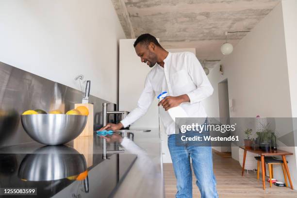 smiling man standing in kitchen of his apartment cleaning countertop - acero inoxidable fotografías e imágenes de stock