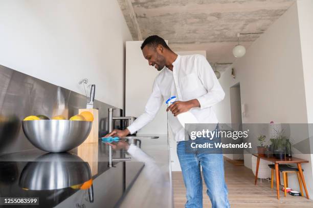 smiling man standing in kitchen of his apartment cleaning countertop - cleaning kitchen stock pictures, royalty-free photos & images