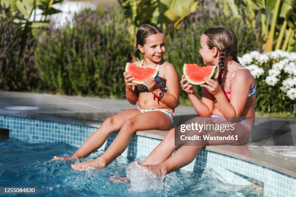 happy twin girls sitting with watermelon slice at poolside on sunny day - twin girls stock pictures, royalty-free photos & images