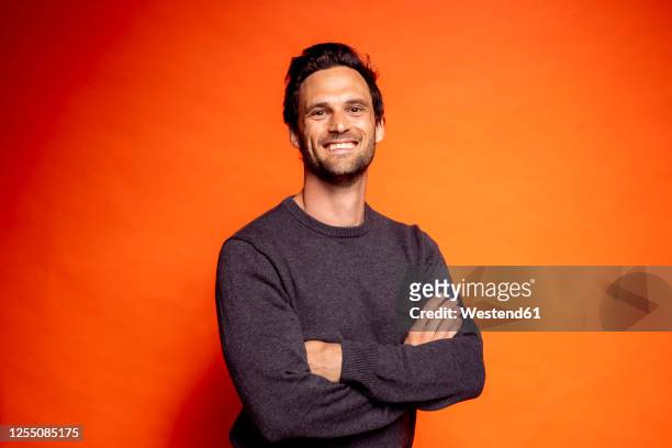 smiling handsome man standing with arms crossed against orange background - oberkörperaufnahme stock-fotos und bilder