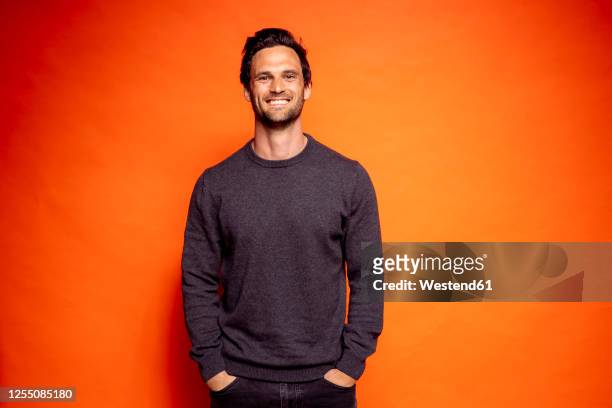happy handsome man with hands in pockets against orange background - three quarter length stock pictures, royalty-free photos & images