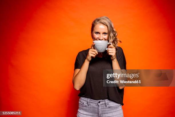 happy woman holding coffee cup with heart shape while standing against orange background - coffee happy stockfoto's en -beelden