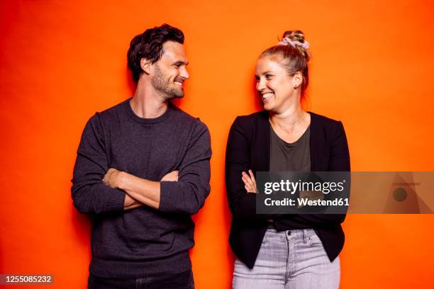 happy mid adult couple looking at each other while standing with arms crossed against orange background - couple sourire studio photos et images de collection