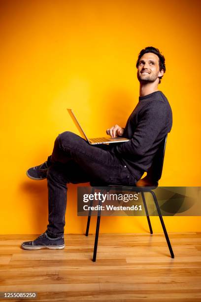 handsome man looking away while sitting with laptop on chair - laptop coloured background stock pictures, royalty-free photos & images