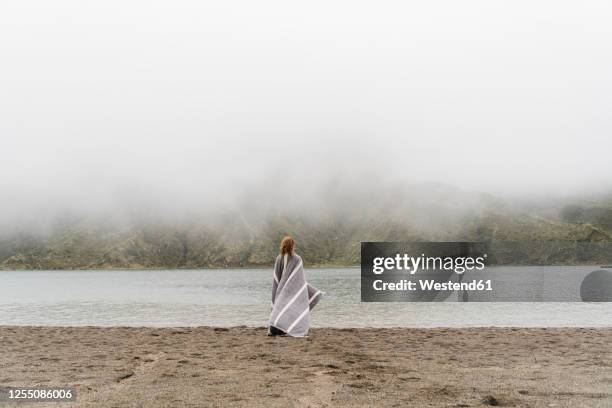 woman wrapped in blanket standing at lakeshore in sao miguel island, azores, portugal - emmitouflé dans une couverture photos et images de collection