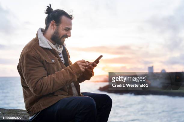 smiling mid adult man using smart phone while sitting against lake at sao miguel island, azores, portugal - volcanic rock bildbanksfoton och bilder
