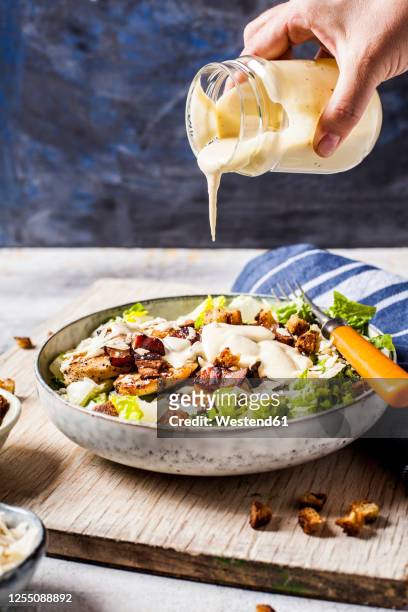 hand of person pouring dressing over bowl of caesar salad with romaine lettuce, parmesancheese, bacon, chicken breast and croutons - sauce stock pictures, royalty-free photos & images