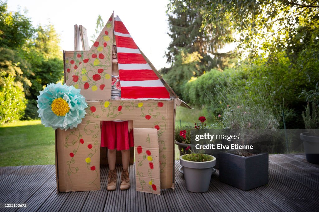Child (6-7) playfully pulling a silly face through the gaps inside a homemade playhouse made from recycled cardboard