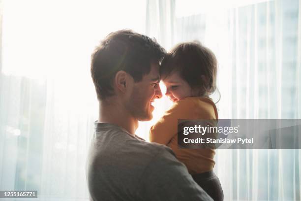 father hugging daughter and smiling against window - lean in collection father stock-fotos und bilder