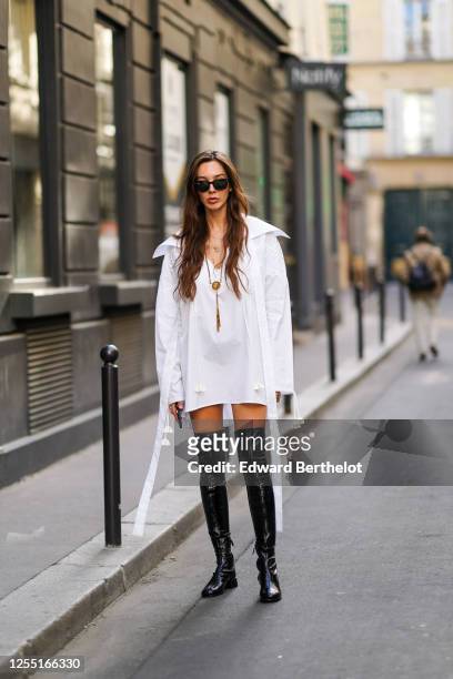 Estelle Chemouny wears Ray-Ban sunglasses, a Charles Jeffrey oversized white long shirt/dress, a golden necklace, Prada thigh high black leather...
