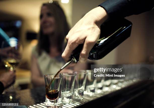 close up of bartender pouring drinks from bottle into a row of shot glasses standing on bar counter, woman sitting in background. - shot glass stock-fotos und bilder