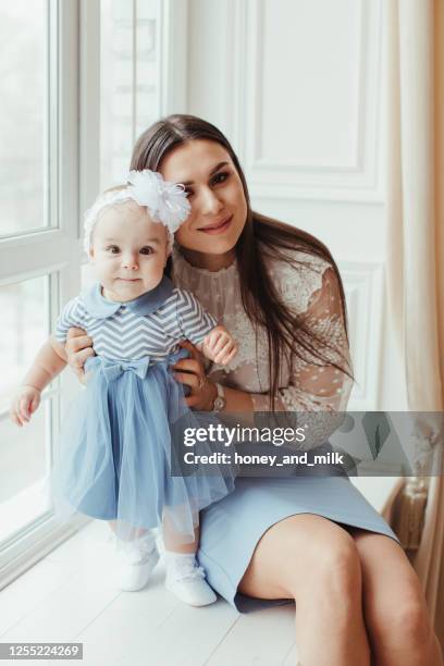 smiling mother sitting by a window with her baby daughter - elegant young woman sitting on window sill stock pictures, royalty-free photos & images