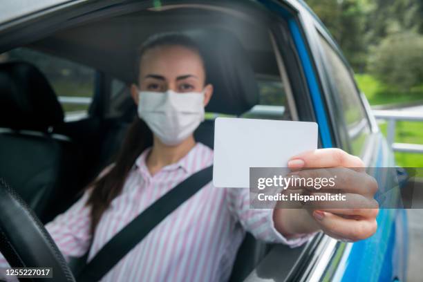 woman driving a car wearing a facemask and holding a card towards the camera - driver license stock pictures, royalty-free photos & images